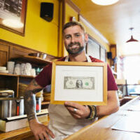 Portrait of male deli owner holding framed dollar bill